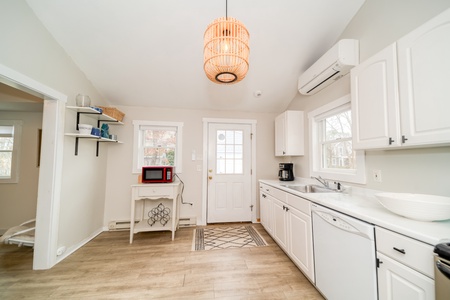 A kitchen with white cabinets, a sink, a dishwasher, a microwave on a small table, and a hanging light fixture. A door with a window and two side windows are in the background.