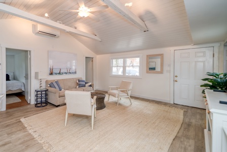 A bright, minimally furnished living room features a beige sofa, two chairs, a wicker table, a large rug, and wall art. There is a door, a window, and visible open doorway to a bedroom.