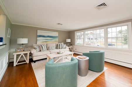 A living room with a beige sofa, patterned pillows, two teal chairs, a white coffee table, and large windows letting in natural light. The walls are painted light grey, and the floor is hardwood.