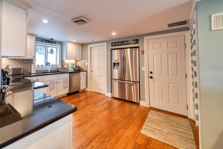 A kitchen with hardwood floors, white cabinetry, stainless steel refrigerator, dishwasher, oven, and a double sink under a window. There are two white doors beside the fridge and a mat on the floor.