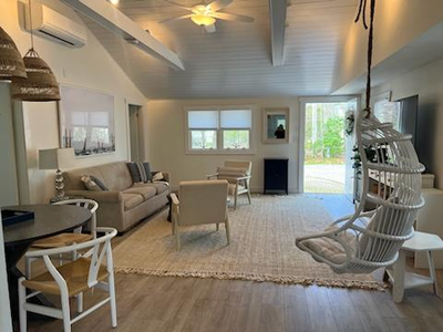 A modern living room with a ceiling fan, cozy sofa, two armchairs, hanging chair, and a rug. The room features large windows, a round dining table, and a view of an outdoor space.