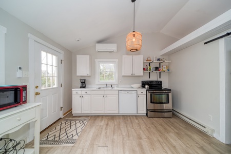 A small, clean kitchen with white cabinets, a stainless steel stove, a dishwasher, and a black microwave on a side table. A hanging light fixture is overhead, and a door with a window is on the left.