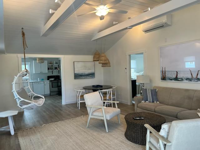 Bright, airy living room with white walls, neutral-colored furniture, a hanging chair, ceiling fan, and open view into a modern kitchen in the background.