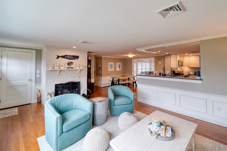 Cozy living room with wooden floors, light blue armchairs, a white coffee table, and a brick fireplace under a fish decoration. An open kitchen with white cabinets is visible in the background.