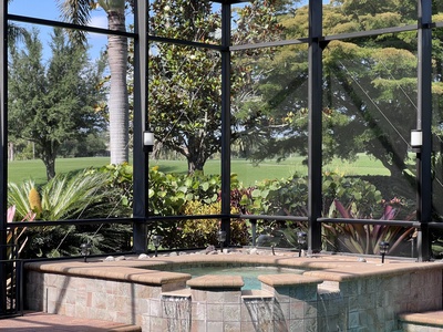 A hot tub with beige tile surrounds is situated within a screened enclosure. Lush greenery, including various trees, shrubs, and plants, can be seen outside the screen.