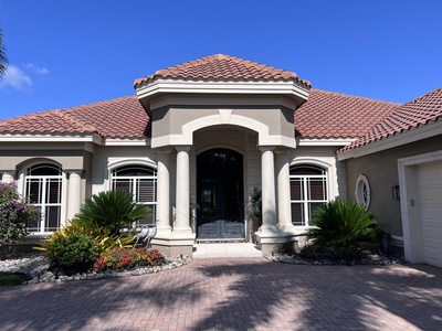 A one-story house with a red-tiled gable roof, beige exterior walls, and a centered grand entrance flanked by columns. The house has shrubs, plants, and a paved driveway in front. The sky is clear and blue.