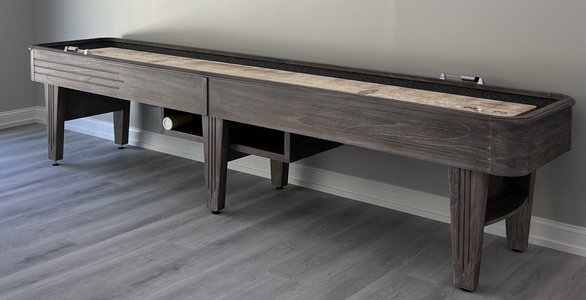 A long, dark wooden shuffleboard table stands against a plain gray wall on a gray wood-patterned floor. There are small storage compartments underneath.