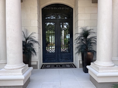 A double-door entrance with ornamental wrought-iron details, flanked by two potted plants. A monogrammed 'B' doormat is placed at the doorway, framed by two large columns.