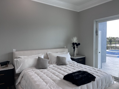 A neatly made bed with white bedding and two grey pillows, beside dark wooden nightstands, in a bedroom with light grey walls and a view of a pool through open sliding glass doors.