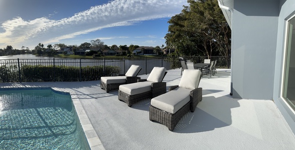 A poolside patio with three cushioned lounge chairs and a table set near a fence overlooking a lake, under a clear blue sky with scattered clouds. Trees and houses are visible in the background.