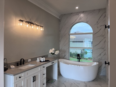 A modern bathroom features a white vanity with a sink, illuminated by a row of bulbs. A freestanding bathtub is set in front of a large arched window with a view of an outdoor garden.