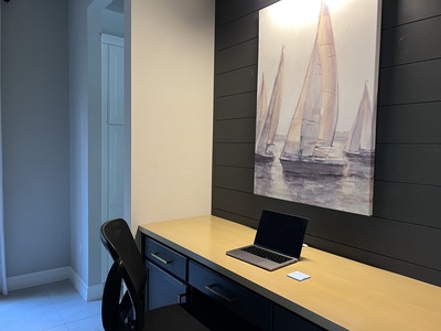 A small home office with a black chair, a wooden desk with drawers, a closed laptop, and a sticky note. A painting of sailboats is mounted on the black wall above the desk.