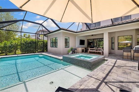 Image of a backyard with a screened-in patio featuring a pool and a connected hot tub. Patio furniture and an outdoor TV are visible, with a garden in the background.