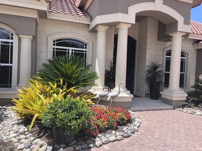 A beige house with a tile roof, flanked by columns. The front yard features colorful flowers, shrubs, rocks, and two decorative bird statues.