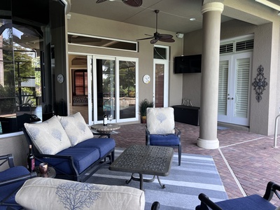 A covered outdoor patio with cushioned chairs and a coffee table on a striped rug. Glass doors and windows provide access to the indoor area, and a wall-mounted TV is beside the doors.