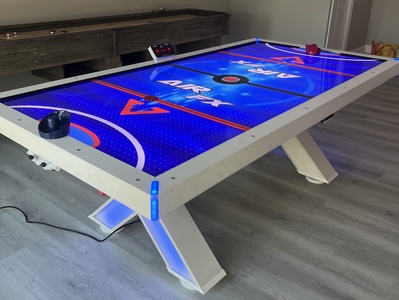 A blue and white air hockey table with a digital scoreboard, two paddles, and a puck, located in a modern room with wooden flooring.