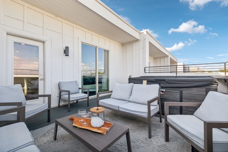 A rooftop patio with cushioned chairs, a wooden coffee table with drinks, and a hot tub. A door and large window are on the adjacent wall. The sky is clear with a few scattered clouds.