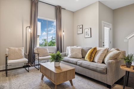 Living room with a gray sofa, white armchairs, a wooden coffee table, and potted plants. A large window with gray curtains lets in natural light. Neutral-toned decor complements the space.