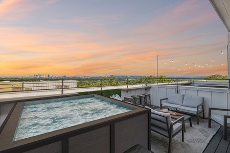 Rooftop terrace with a hot tub, outdoor seating area, string lights, and a city skyline view at sunset.