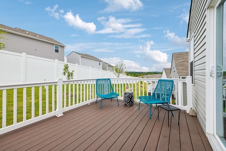 A wooden deck with two blue chairs and a small table overlooks a grassy backyard, surrounded by white fences and neighboring houses under a blue sky with clouds.