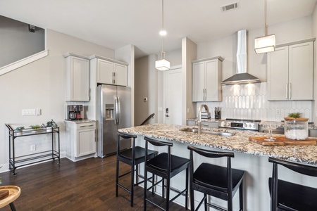 Modern kitchen with white cabinets, stainless steel appliances, granite countertops, black bar stools at an island, a tile backsplash, and pendant lights. Wooden floor and staircase to the left.