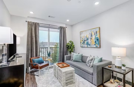 A modern living room with a gray sofa, two ottomans, a leather chair, a large window with curtains, wall artwork, and various decor items. The room has hardwood flooring and recessed lighting.