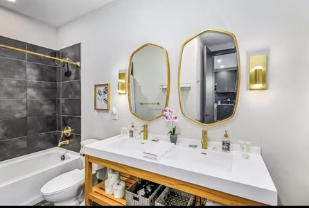 A modern bathroom with a double sink vanity, two oval mirrors, a bathtub with dark grey tiles, and gold fixtures. A small orchid plant and toiletries are on the countertop.