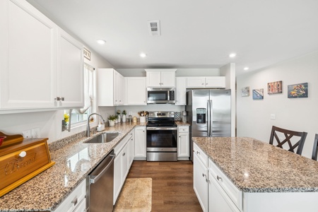 A modern kitchen with white cabinetry, stainless steel appliances, granite countertops, and a kitchen island with bar seating. Artworks adorn the adjacent dining area wall.