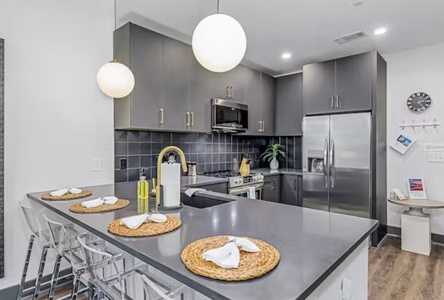 Modern kitchen with dark gray cabinets, stainless steel appliances, and a central island with four place settings. Pendant lights hang above the island, and a clock is on the wall.