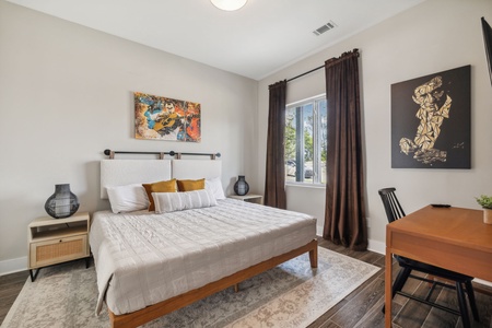 A bedroom with a large bed, white linens, and a wooden frame. It has two bedside tables, wall art, a window with dark curtains, and a wooden desk with a black chair. Rug covers the floor beneath the bed.