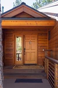 Wooden cabin entrance with a wooden door and side window, surrounded by trees. A small porch and railing lead to the door.