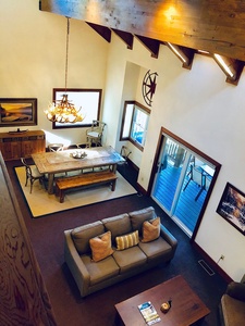 Aerial view of a living room with a sofa, coffee table, dining table, chandelier, and patio doors leading to a deck. Walls are decorated with art, and the ceiling has exposed beams.