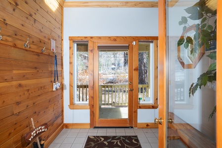 Wood-paneled entryway with glass door leading outside. Hooks on the left wall. Brown mat on tiled floor, plants on the right.