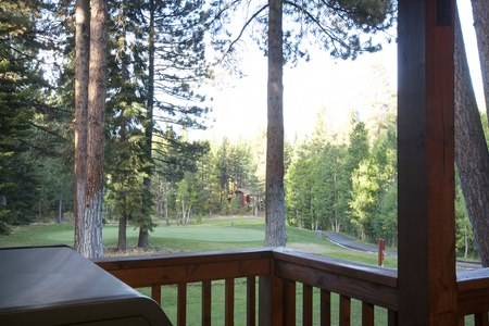 A wooden deck overlooks a lush, green golf course surrounded by tall trees. A grill is partially visible in the foreground.