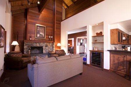 A cozy living room with a stone fireplace, wooden walls, two chairs, a sofa, and a small kitchen area with a wine cooler. Warm lighting and artwork enhance the ambiance.