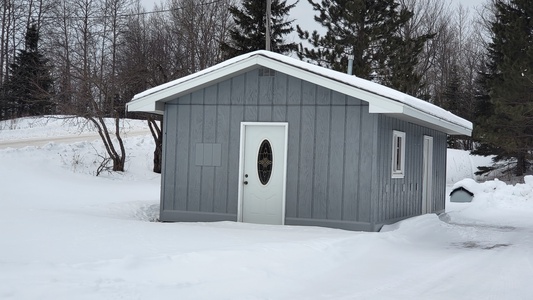 A small, gray wooden cabin with a white door is surrounded by snow. Snow-covered trees and a road are visible in the background. The sky is overcast.