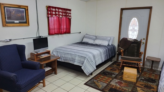 A small room with a bed, armchair, rocking chair, side table with a small TV, a door with a decorative glass, and a window with a red checkered curtain. A framed picture hangs on the wall.