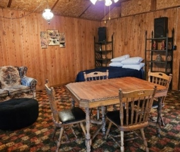 Room with wooden walls and ceiling, a table with four chairs, a bed with pillows, a patterned sofa, shelves, and various decorations.