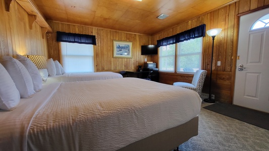 A cozy hotel room with wood-paneled walls, two double beds, a desk with a chair, a TV, and large windows adorned with navy curtains. A floor lamp stands near the door.