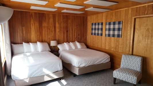 A hotel room with wooden paneling features two double beds with white bedding, a patterned chair, a reading lamp, and two checkered wall hangings.