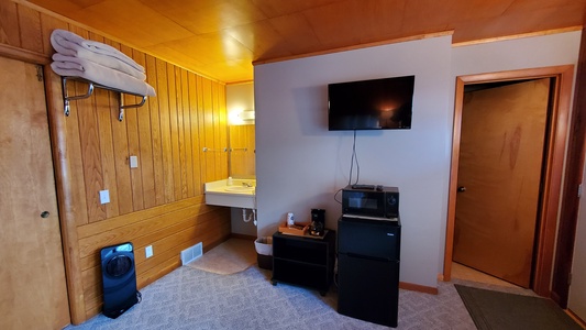 A small room with wood-paneled walls, a wall-mounted sink, a flat-screen TV, a mini-fridge, a microwave, a coffee maker, a folded towel rack, and a portable heater. Door is visible on the right.