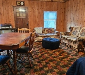 A wood-paneled room features a dining table with chairs, a floral-patterned sofa and loveseat, a blue ottoman, a window with blinds, and vintage electronics on a cabinet. The carpet is multicolored.