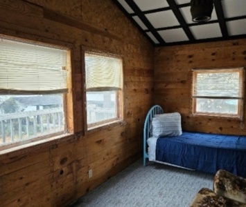 A small room with wooden walls, featuring three windows, a single bed with a blue blanket and pillows, and a carpeted floor.