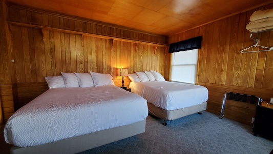 A wooden bedroom features two neatly made beds with white linens, a small bedside table with a lamp, folded towels on a wooden shelf, and a window with a blue curtain.