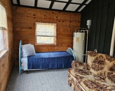 A small room with wood-paneled walls featuring a single bed with a blue bedspread, a floral-patterned sofa, and a water heater. There are two windows, one above the bed, allowing natural light into the room.