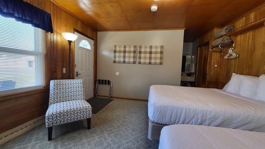 A cozy bedroom with two beds, a patterned armchair, a small lamp, and wall art. The room has wood-paneled walls, a window with blinds, and an open closet area with hangers and folded towels.