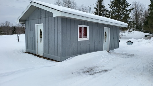 A small gray wooden building with two white doors and a window is surrounded by snow on a winter day.