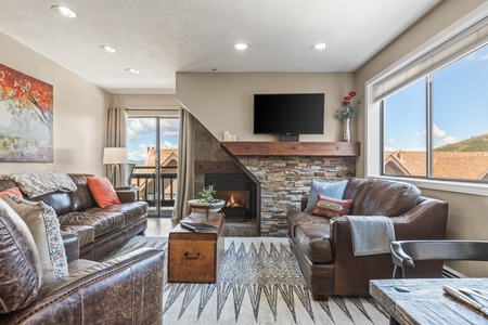 A living room features leather sofas, a stone fireplace topped with a flat-screen TV, large windows, and a rug with geometric patterns.