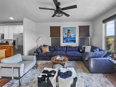 A living room with a large blue sectional sofa, a white armchair, two black and white ottomans, a round wooden coffee table, and a ceiling fan. A kitchen is visible in the background.