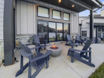 A modern outdoor patio featuring six dark Adirondack chairs surrounding a lit fire pit, adjacent to a building with large windows and a covered roof.